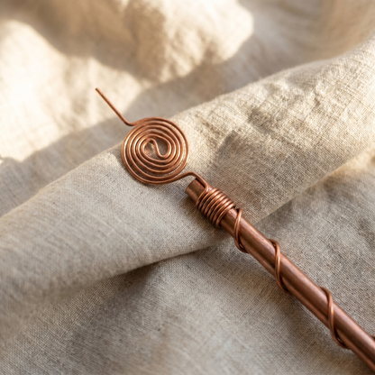A single Henri's Plant-Boosting copper garden stake lying diagonally on softly creased oatmeal linen cloth, soft warm morning light revealing the helical wrap and spiral coil head detail.